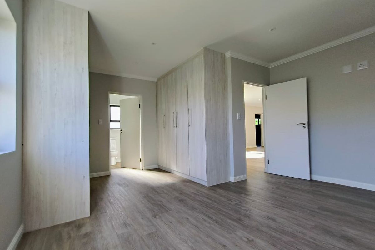 A bright, modern bedroom featuring light wood-grain built-in cupboards, neutral grey walls, and durable wood-look laminate flooring, with an en-suite bathroom access.