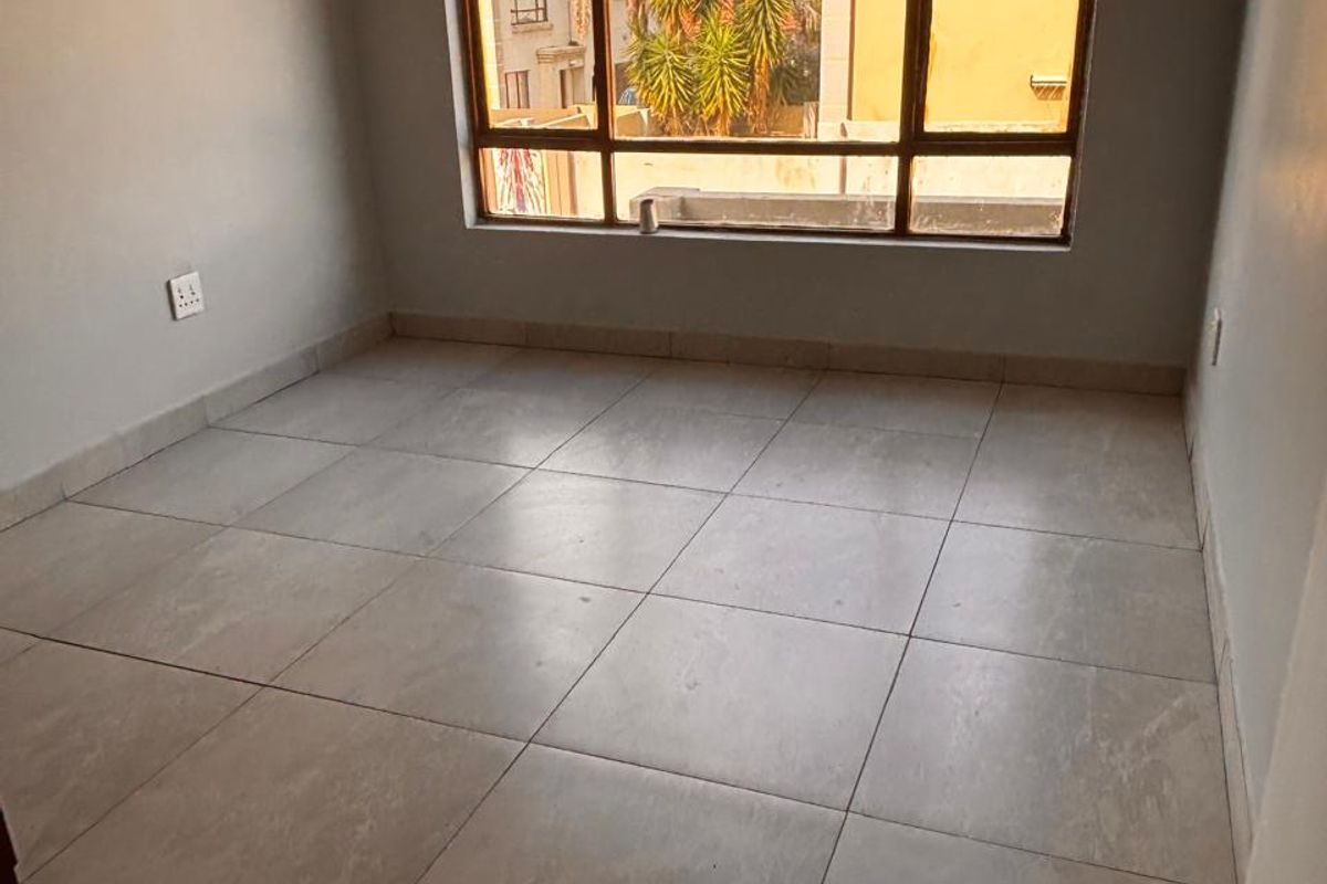 A bright, empty bedroom featuring large windows with neighborhood views and neutral tiled flooring.