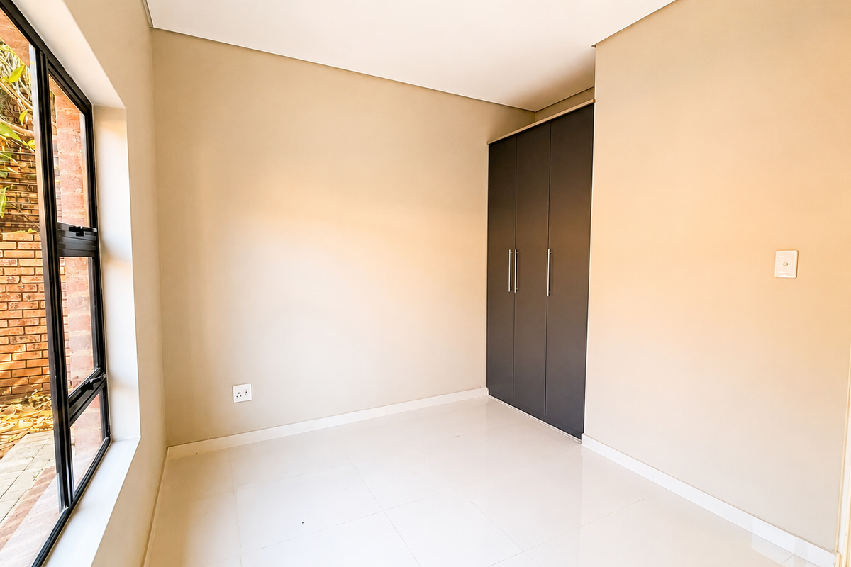 A bright, empty bedroom featuring neutral tiled flooring, smooth walls, and a modern built-in wardrobe with dark grey doors.