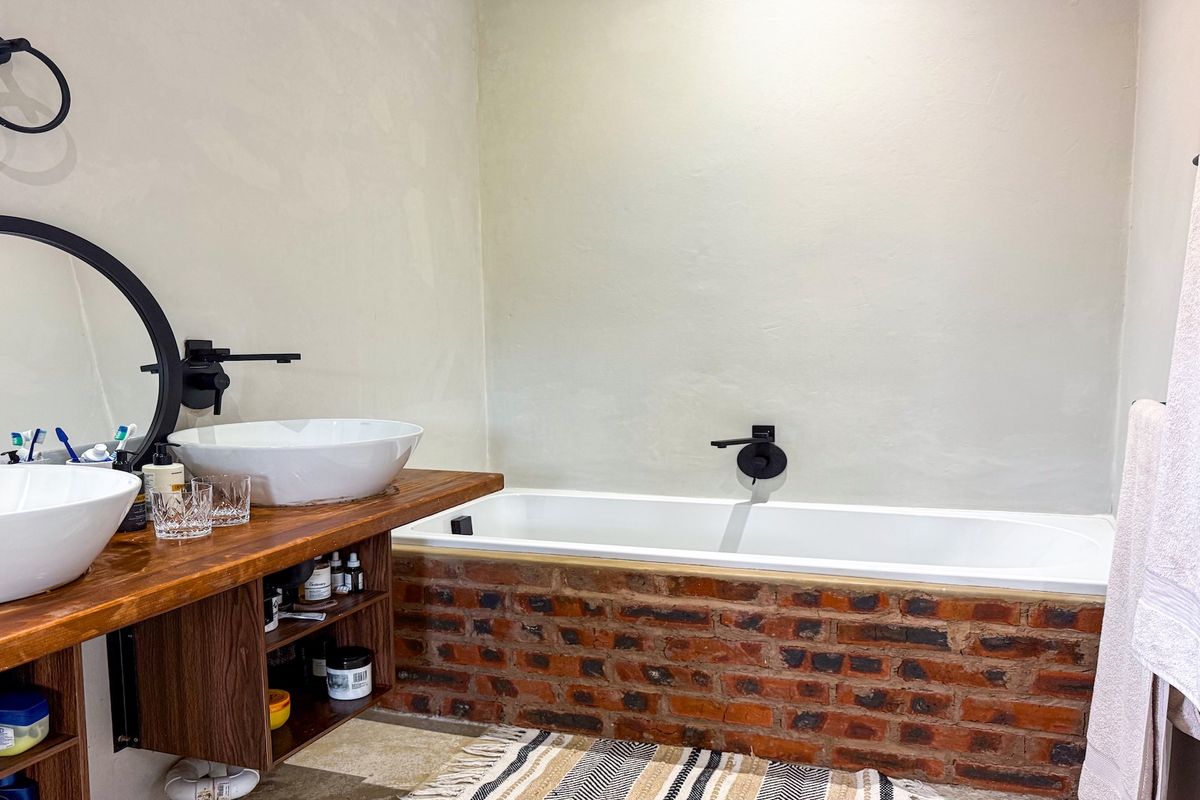Modern bathroom featuring a custom timber vanity with dual vessel sinks, matte black fixtures, and a unique exposed brick bathtub surround.