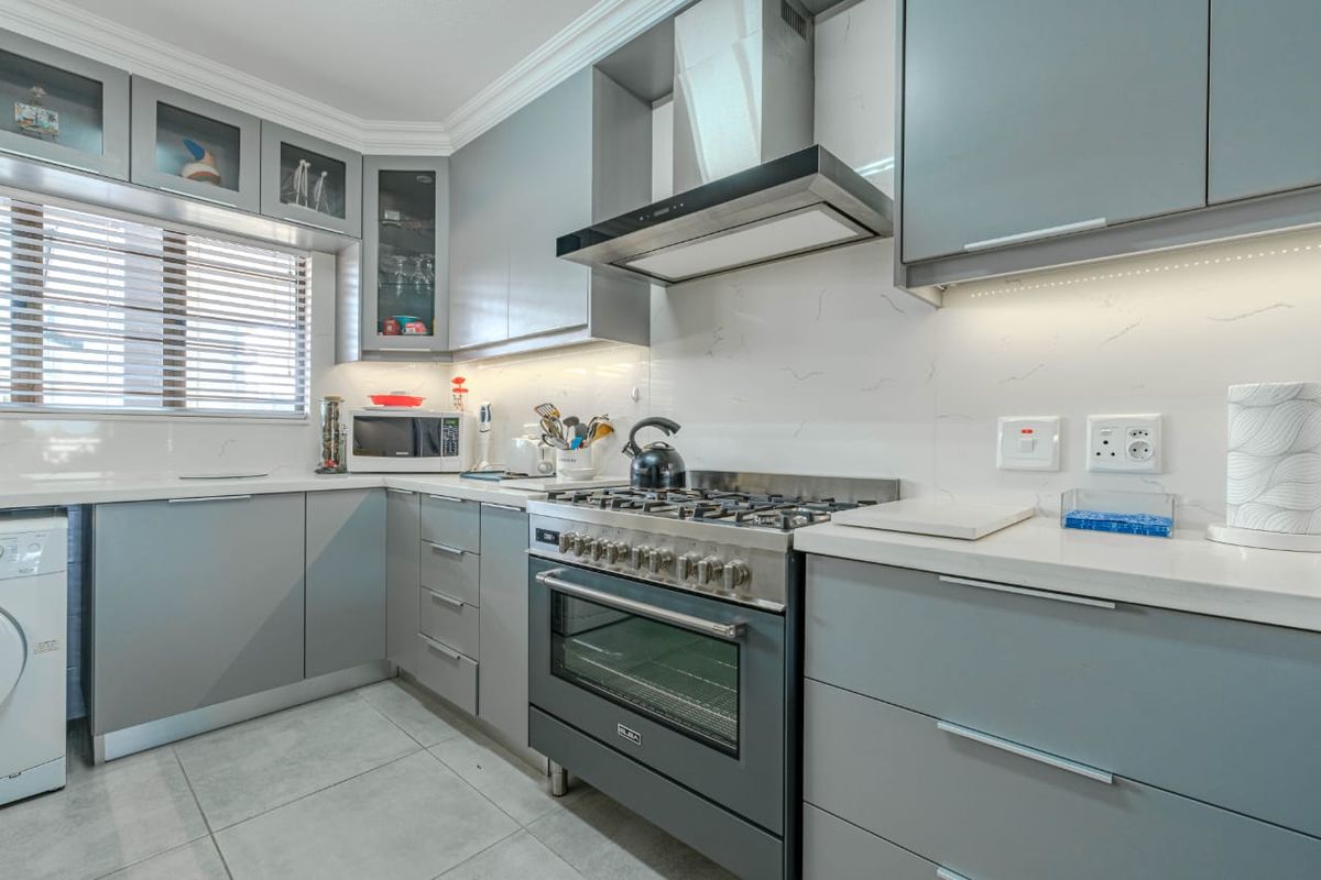 Modern kitchen featuring grey cabinetry, stone countertops, marble-look backsplash, stainless steel gas stove with extractor fan, and integrated under-counter laundry appliance space.