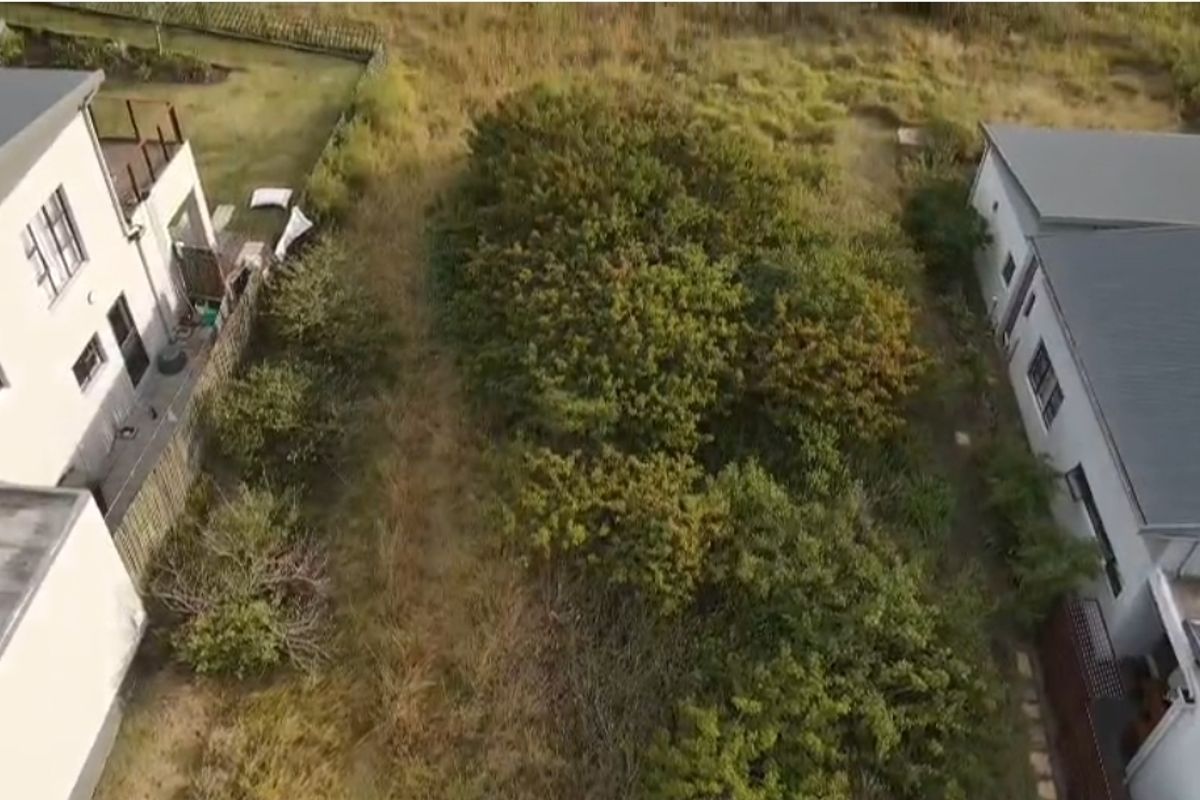 An aerial view of a vacant residential plot situated between two existing properties, featuring natural vegetation and grass cover.