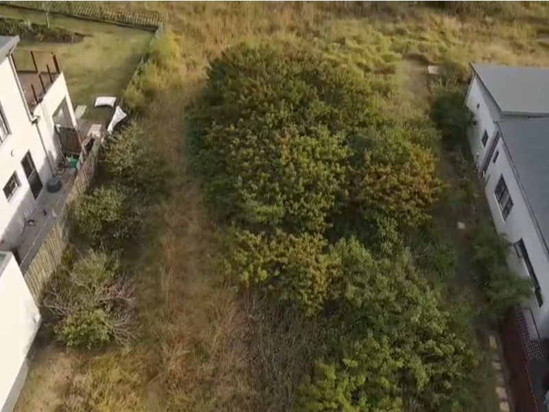 An aerial view of a vacant residential plot situated between two existing properties, featuring natural vegetation and grass cover.