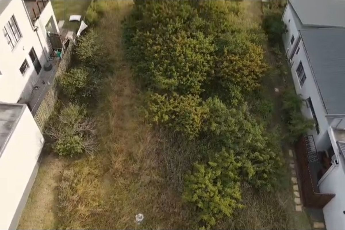 An aerial view of a vacant residential plot situated between existing neighboring properties, featuring overgrown vegetation and natural ground cover.