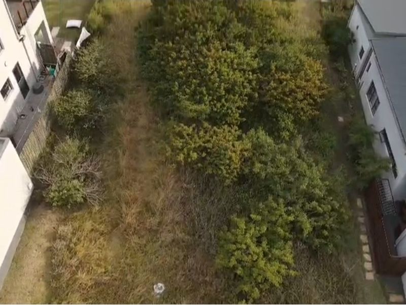 An aerial view of a vacant residential plot situated between existing neighboring properties, featuring overgrown vegetation and natural ground cover.