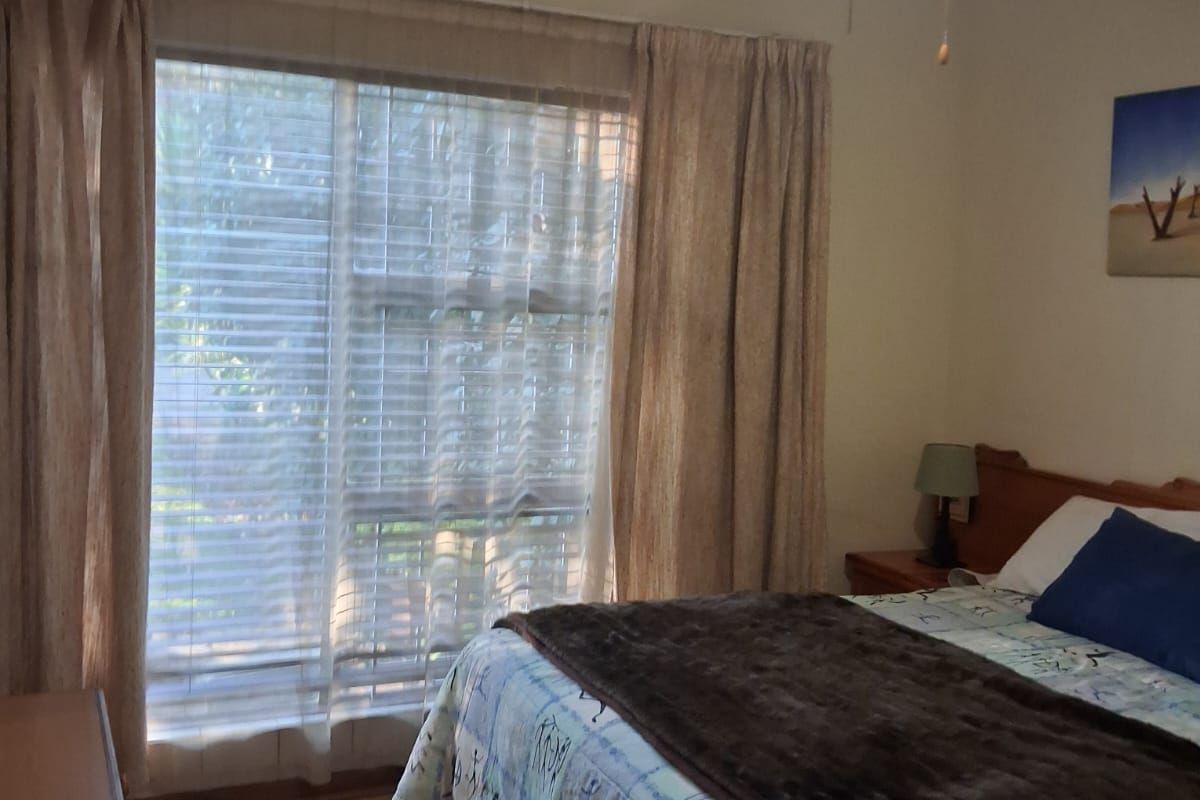 A bedroom featuring wood-look flooring, a ceiling fan, and a large window with blinds and curtains.
