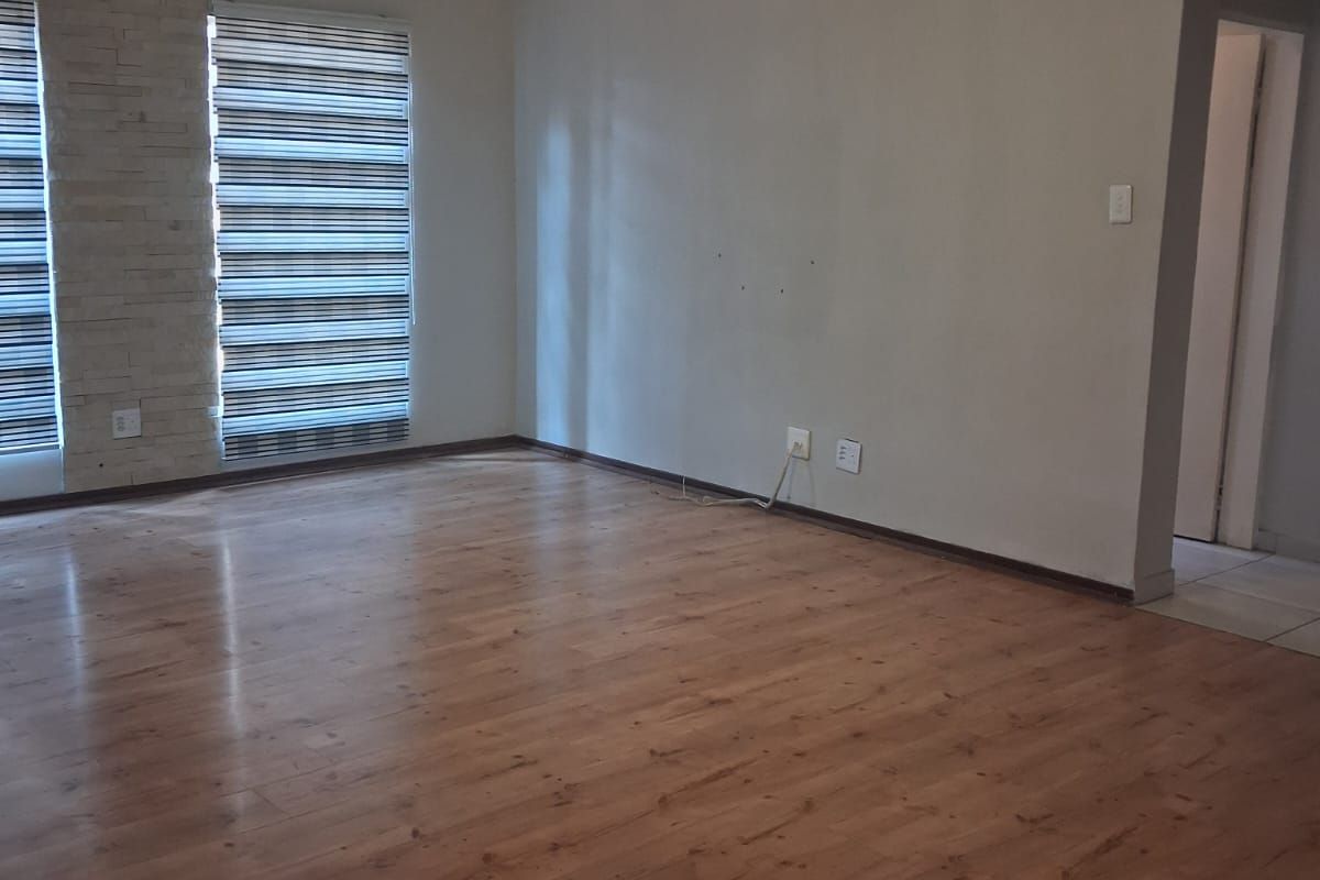 A spacious, empty living room featuring warm-toned laminate flooring, decorative stone-cladding accents around the windows, and modern zebra-style blinds.