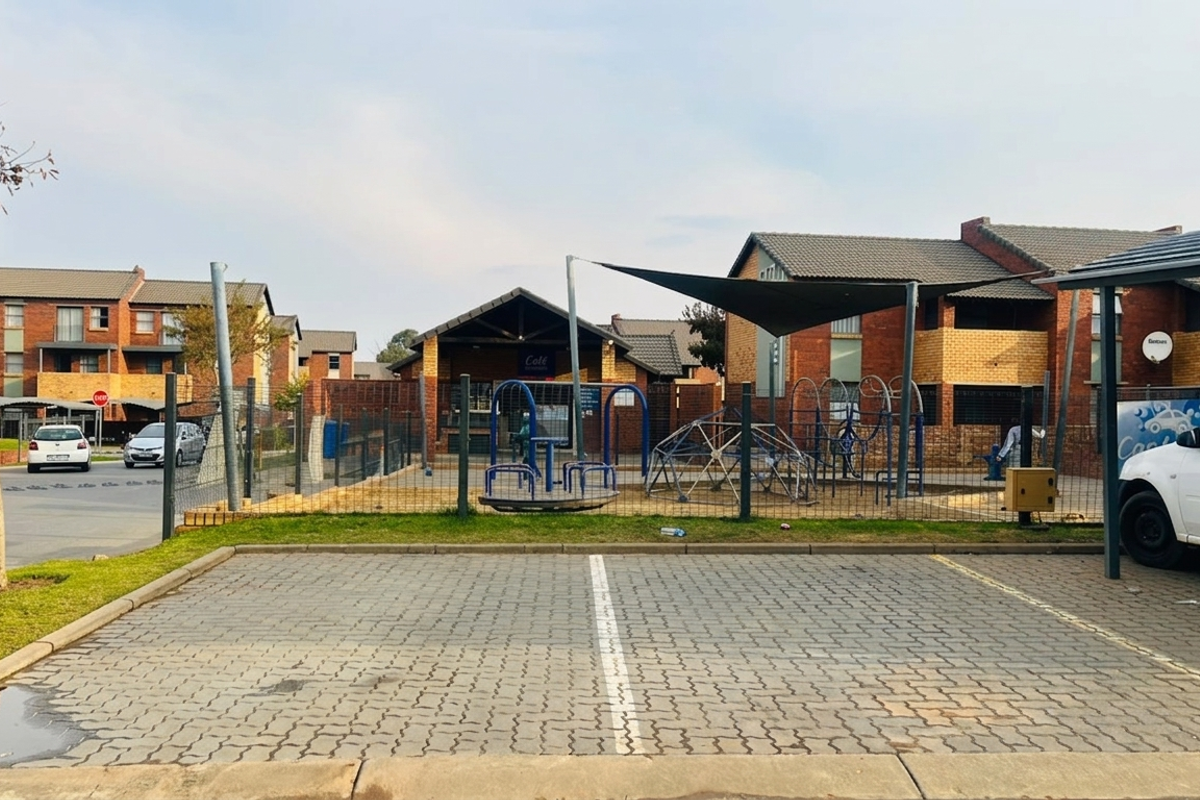 Exterior view of a residential complex featuring a paved parking area, a fenced communal playground with shade sails, and multi-story brick apartment buildings.