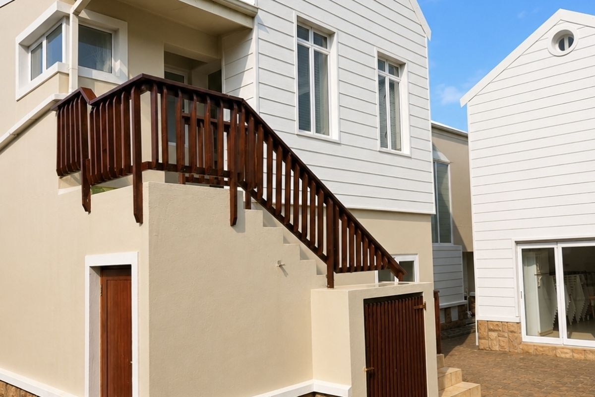 A well-maintained exterior rear view of the house featuring white weatherboard cladding, stone base detailing, a wooden staircase, and a paved courtyard area.