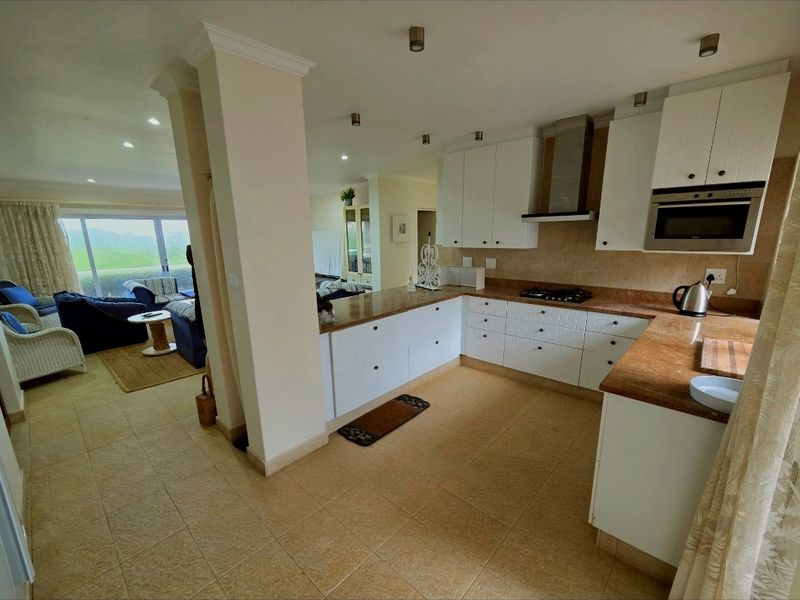 Open-plan kitchen featuring white cabinetry, stone-look countertops, built-in oven, and extractor fan, flowing into a bright living area with large windows.