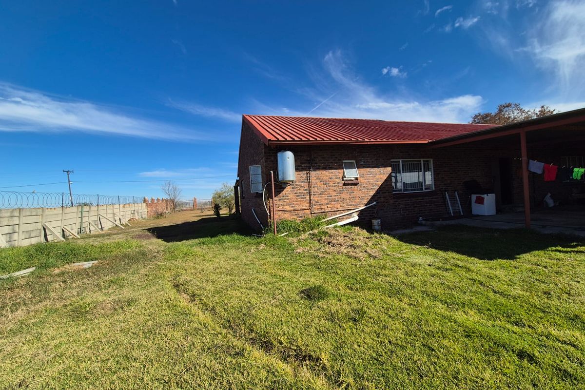 Rear view of a brick house featuring a red metal roof, covered patio area, and a large, unmaintained garden with perimeter walling.