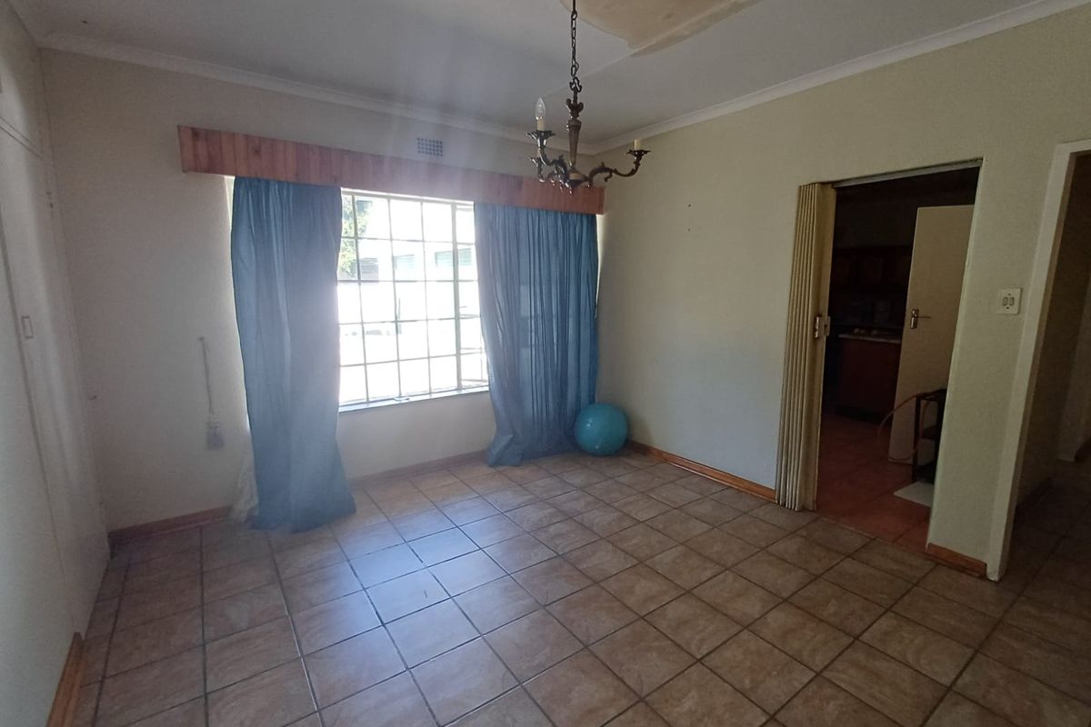 A spacious living area featuring tiled flooring, a large window with wooden pelmet, and a ceiling showing signs of water damage.