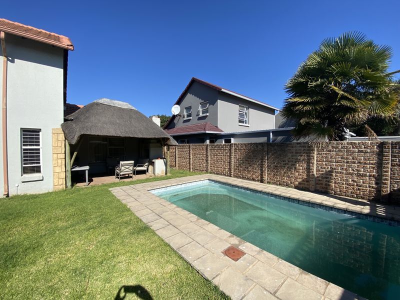 Rear garden featuring a rectangular swimming pool, paved surround, and a covered thatch-roof patio area enclosed by a brick boundary wall.