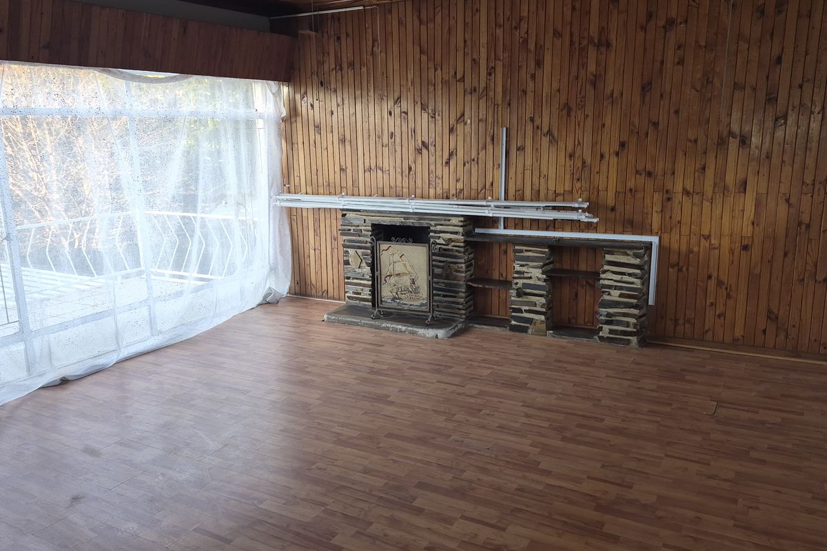 A spacious living room featuring wood-paneled walls, a stone-clad fireplace, and large windows providing natural light and access to an outdoor balcony area.