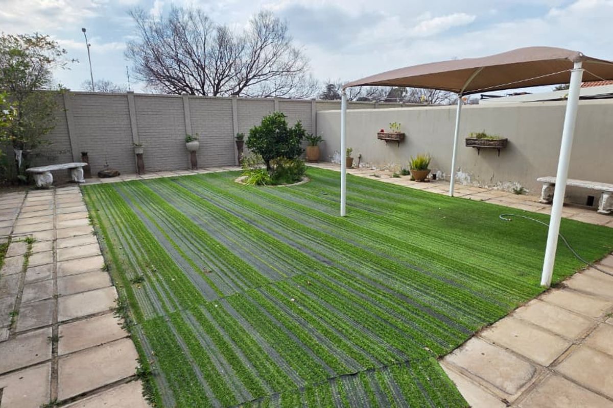 A private rear garden featuring a central artificial turf area, paved surrounds, and a fixed shade structure against a high boundary wall.