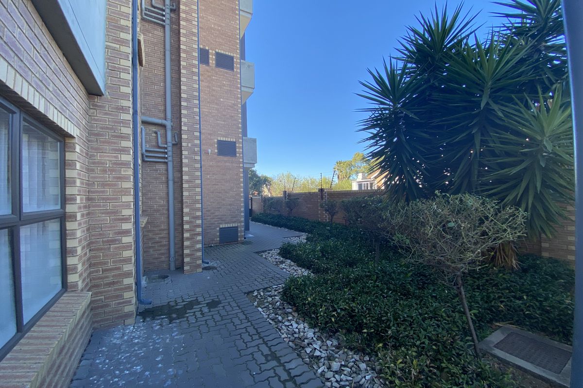 A view of the apartment building's exterior side path, featuring brick facade, paved walkway, and landscaped garden area.