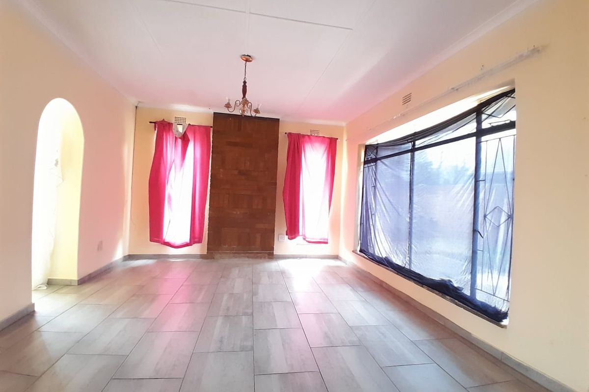 A spacious, light-filled living room featuring wood-look tile flooring, a central brick feature wall, and large windows with security bars.