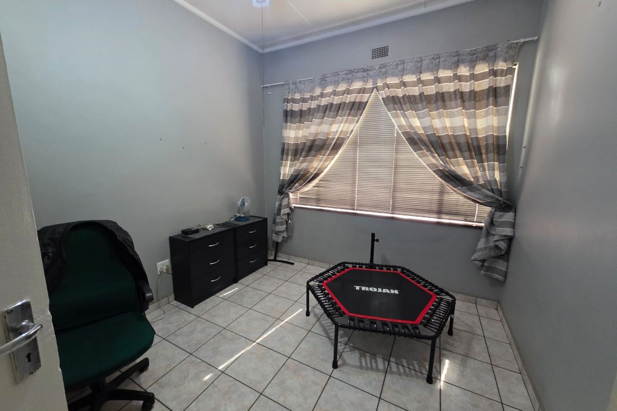 A functional bedroom featuring tiled flooring, neutral grey walls, and a window with fitted blinds and curtains.