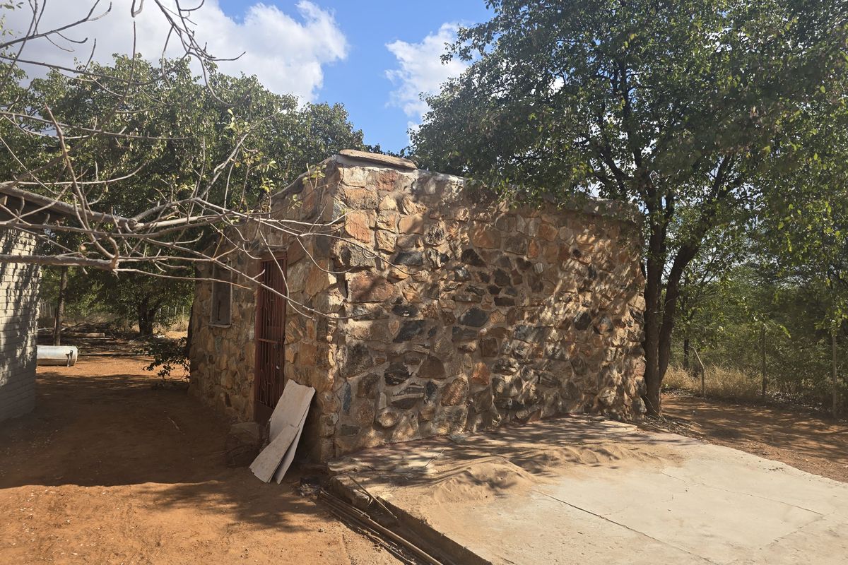 A rustic stone-walled outbuilding on a farm property, featuring a concrete foundation and metal security door, set within a natural landscape.