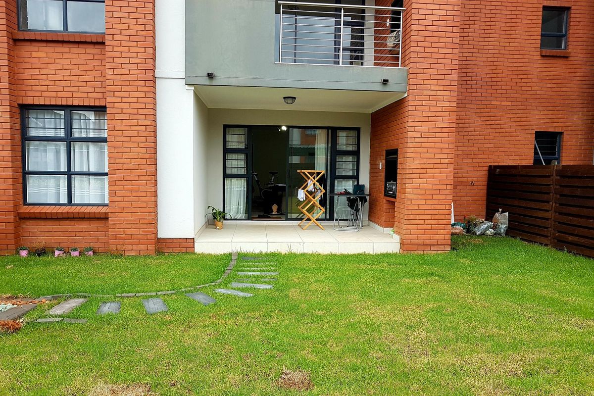 Rear view of the townhouse featuring a private garden, tiled patio with built-in braai, and an upper-level balcony with stainless steel railing.