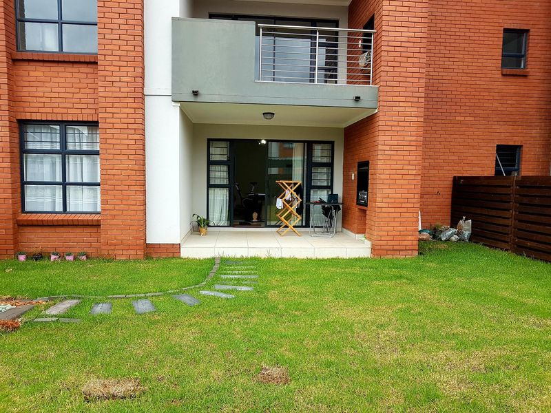Rear view of the townhouse featuring a private garden, tiled patio with built-in braai, and an upper-level balcony with stainless steel railing.