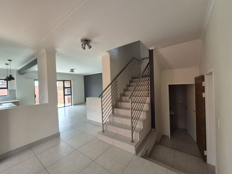 An open-plan living area featuring a modern staircase with stainless steel railings, tiled flooring, and a view into the adjacent kitchen space.
