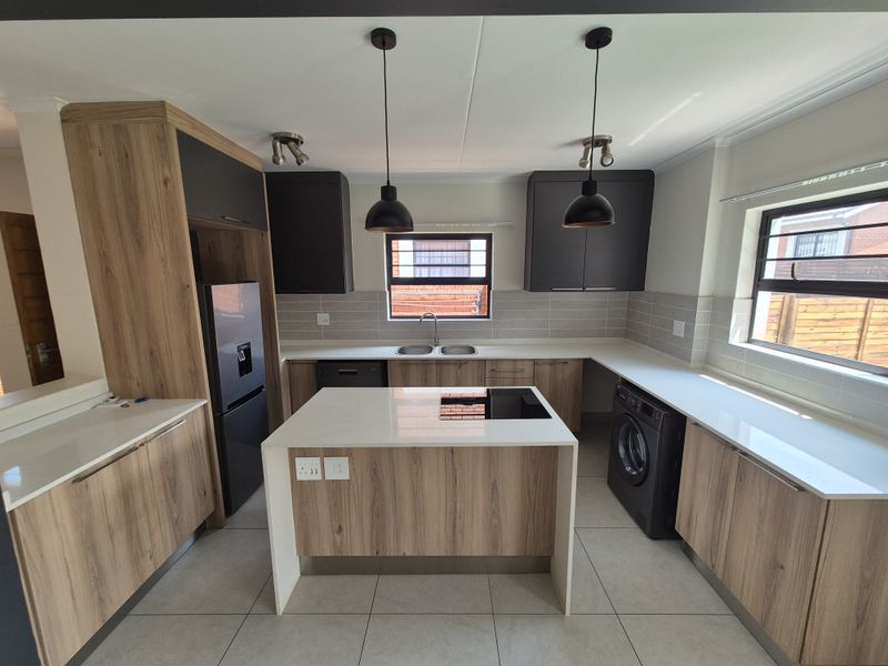 Modern kitchen featuring wood-grain cabinetry, quartz countertops, a central island with integrated hob, double basin sink, and contemporary pendant lighting.