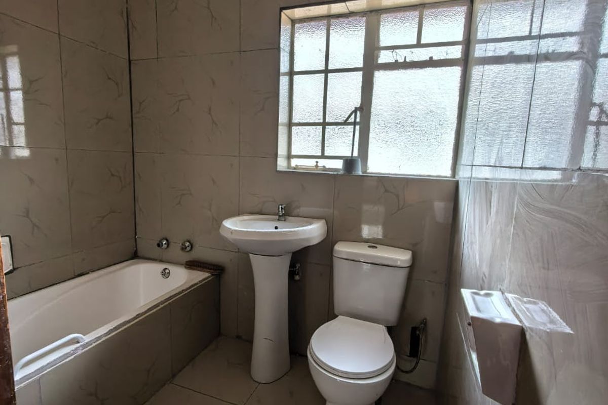 A functional bathroom featuring a built-in bathtub, pedestal basin, and toilet, finished with full-height wall tiling and a window for natural light.