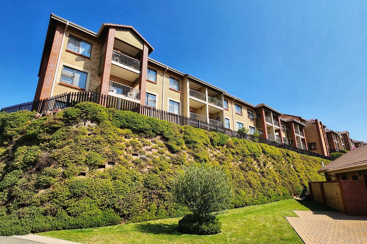 Exterior view of a multi-story apartment complex featuring brick facades, private balconies, and a well-maintained landscaped retaining wall and garden area.