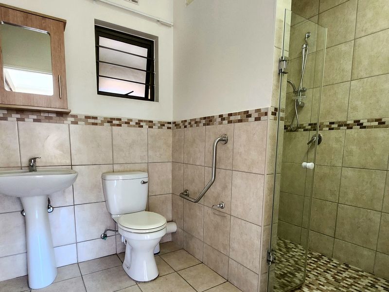 A functional bathroom featuring a pedestal basin, toilet with grab rail, and a glass-enclosed shower with neutral tiled finishes.