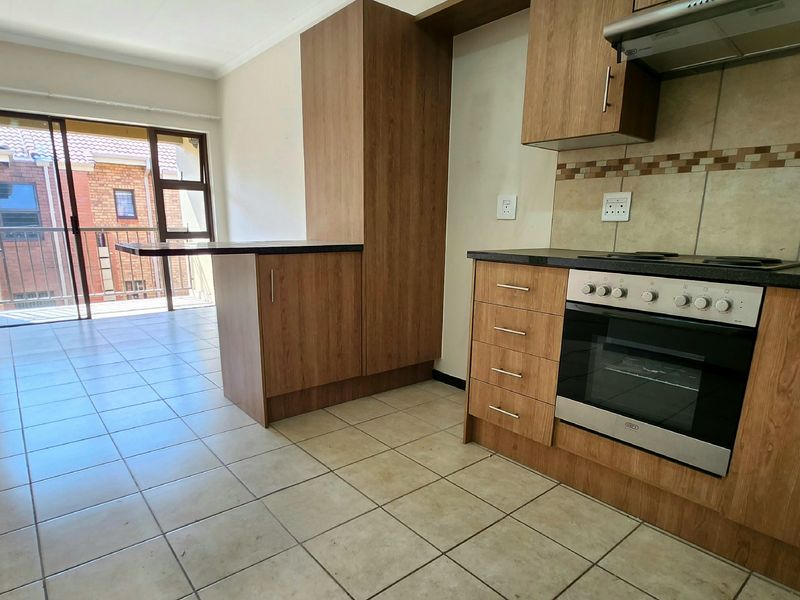 Open-plan kitchen and living area featuring wood-finish cabinetry, built-in oven and hob, tiled flooring, and access to a balcony through sliding glass doors.