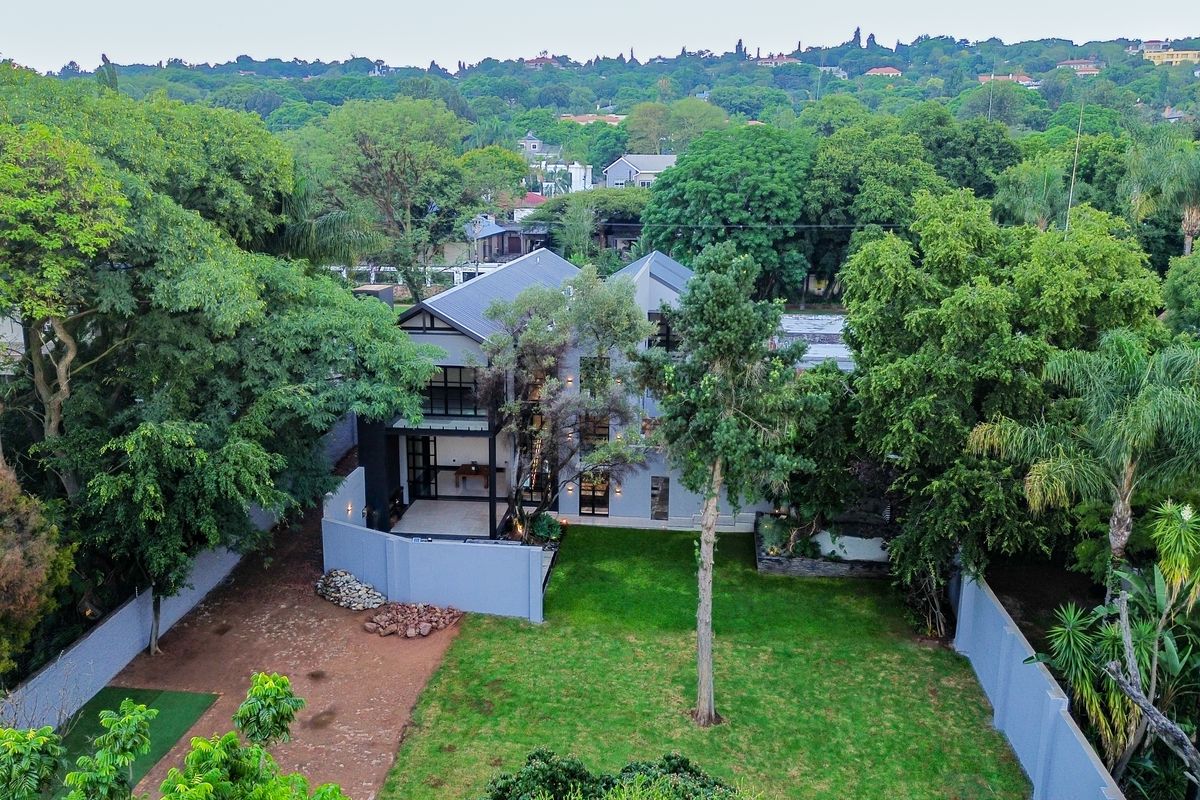 Aerial view of a modern multi-story residence featuring a spacious backyard, lush surrounding greenery, and a paved driveway area.