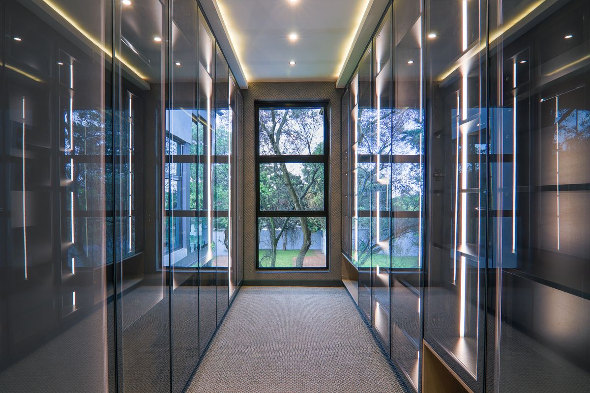 A modern walk-in dressing room featuring floor-to-ceiling glass-fronted cabinetry with integrated LED lighting and a large window overlooking the garden.