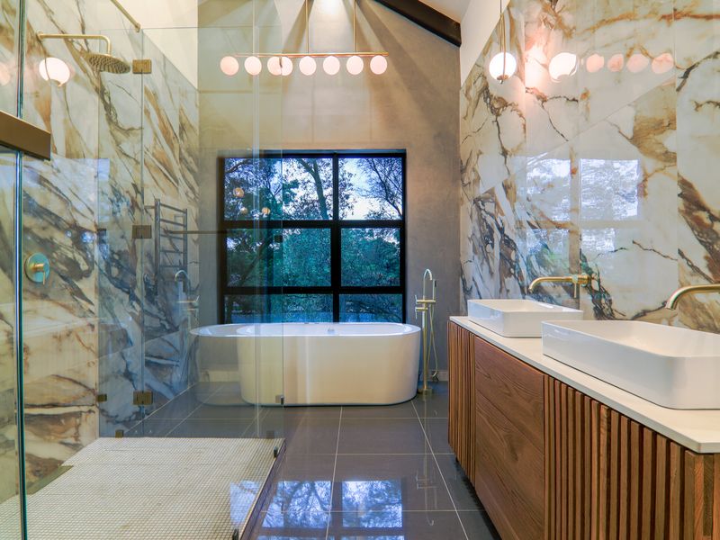 Modern luxury bathroom featuring a freestanding bathtub, glass-enclosed walk-in shower, dual vanity with marble-style wall tiling, and large window with garden views.