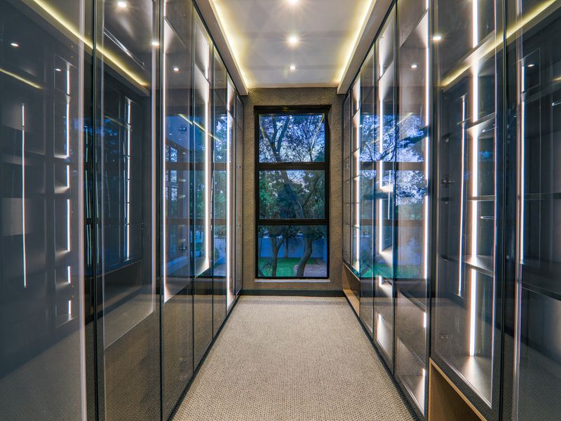 A modern walk-in dressing room featuring floor-to-ceiling glass-fronted cabinetry with integrated LED lighting and a large window overlooking the garden.