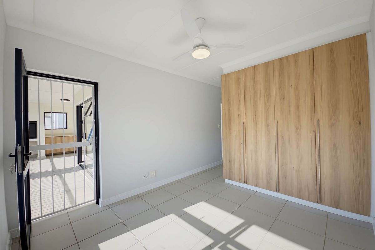 A bright, modern bedroom featuring light-toned floor tiles, built-in wooden cabinetry, and a ceiling fan, with a security gate leading to an adjacent space.