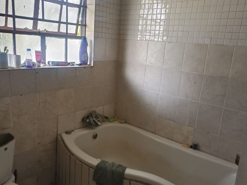 A dated bathroom featuring a built-in bathtub with tiled surround, tiled walls, and a steel frame window. The space requires cosmetic updates and maintenance.