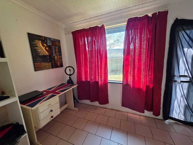 A functional bedroom or office space featuring tiled flooring, built-in shelving, and large windows with red curtains.