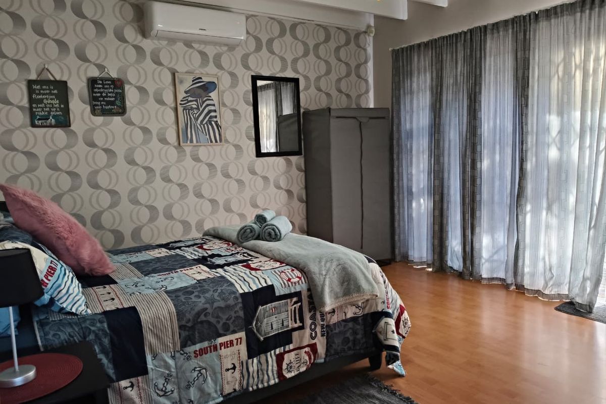 A bedroom featuring wood-look flooring, patterned wallpaper, exposed ceiling beams, and a wall-mounted air conditioning unit with access to a patio through glass doors.