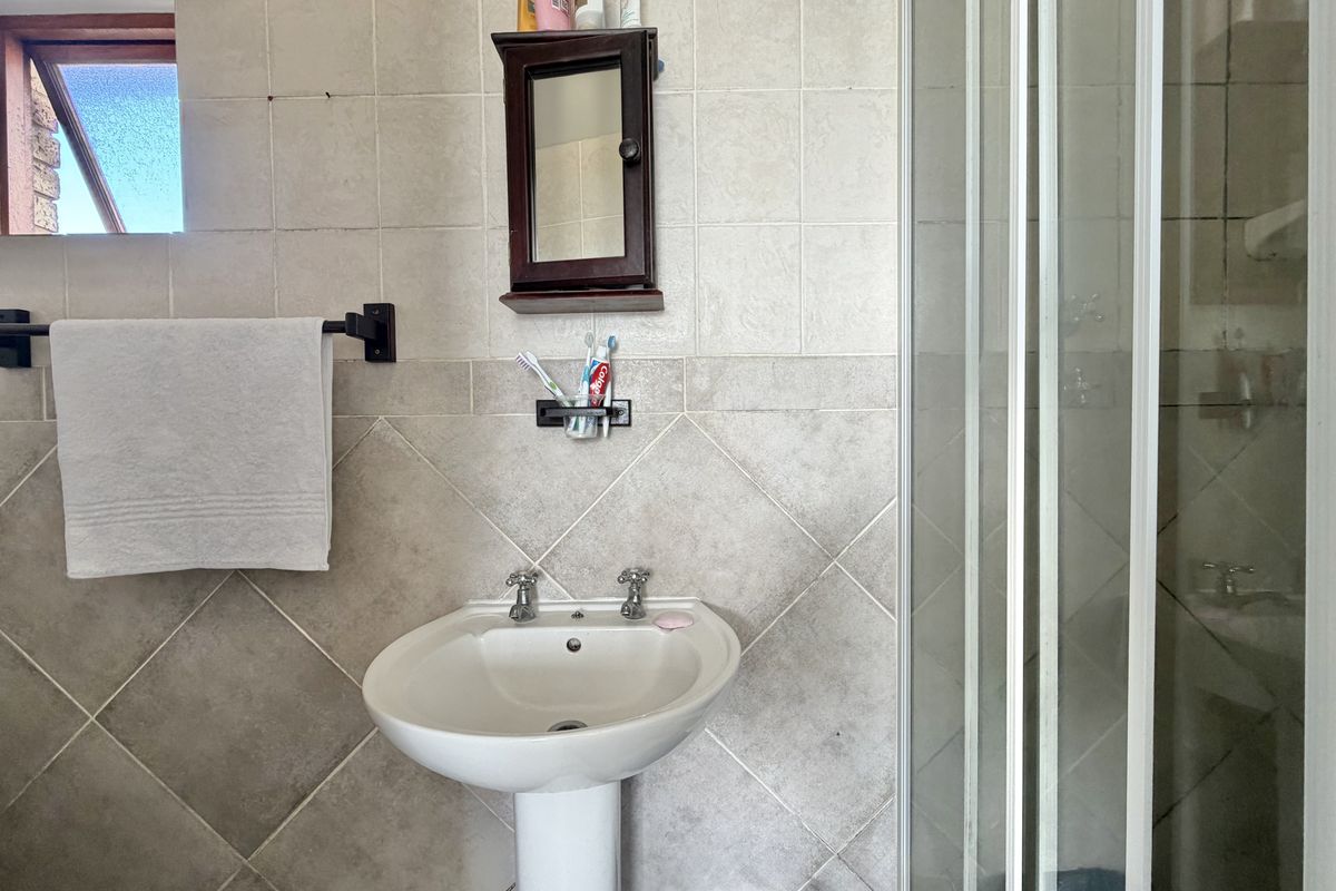 A functional bathroom featuring a pedestal basin, tiled walls, and a glass-enclosed shower stall with natural light from a high-set window.
