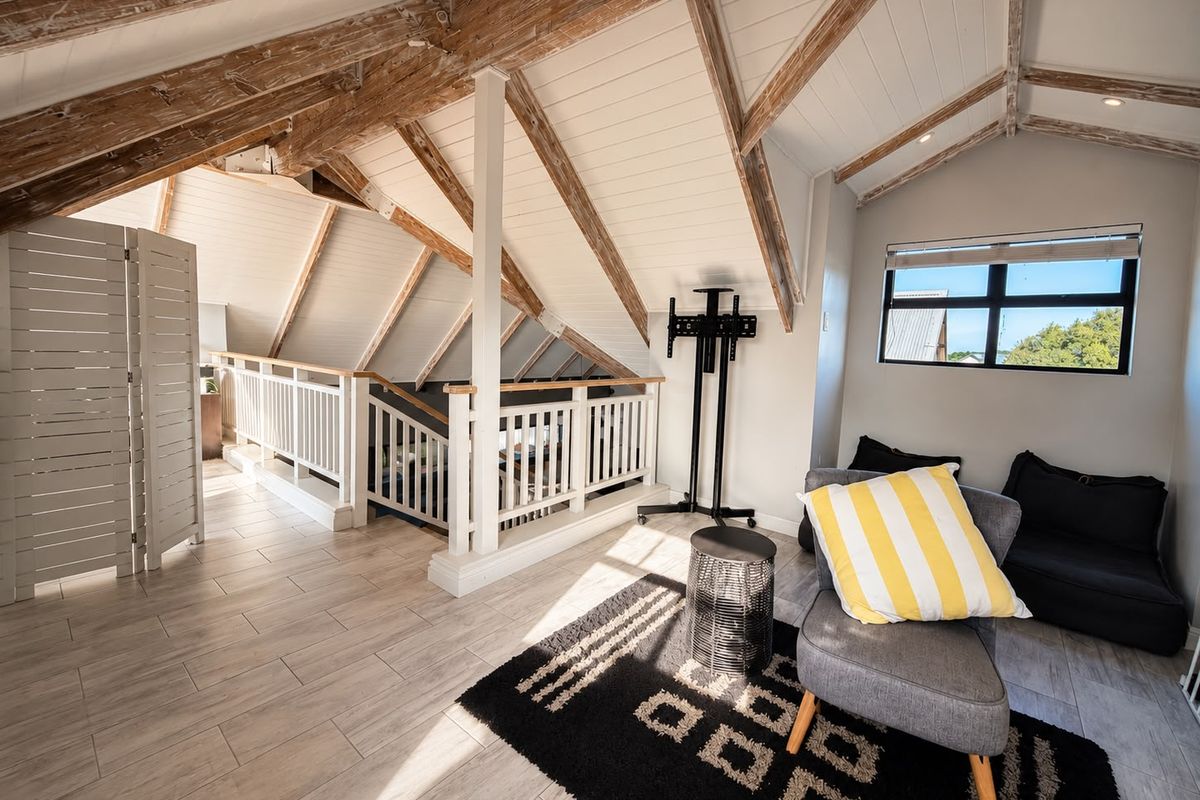 A bright loft living area featuring exposed timber roof trusses, vaulted ceilings, tiled flooring, and a mezzanine-style balustrade overlooking the lower level.