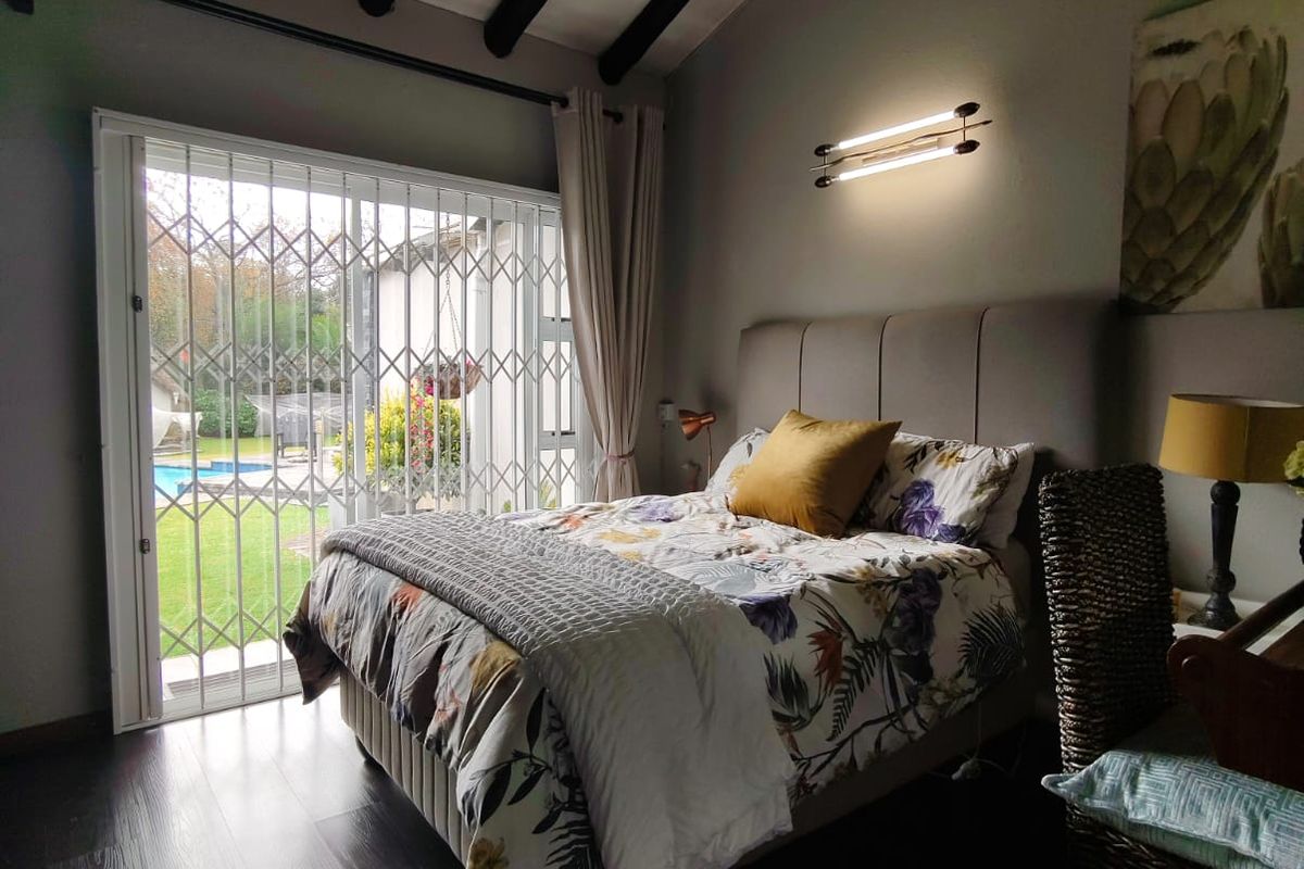 A well-maintained bedroom featuring exposed timber ceiling beams, a ceiling fan, and sliding glass doors with security gates leading to the garden and pool.