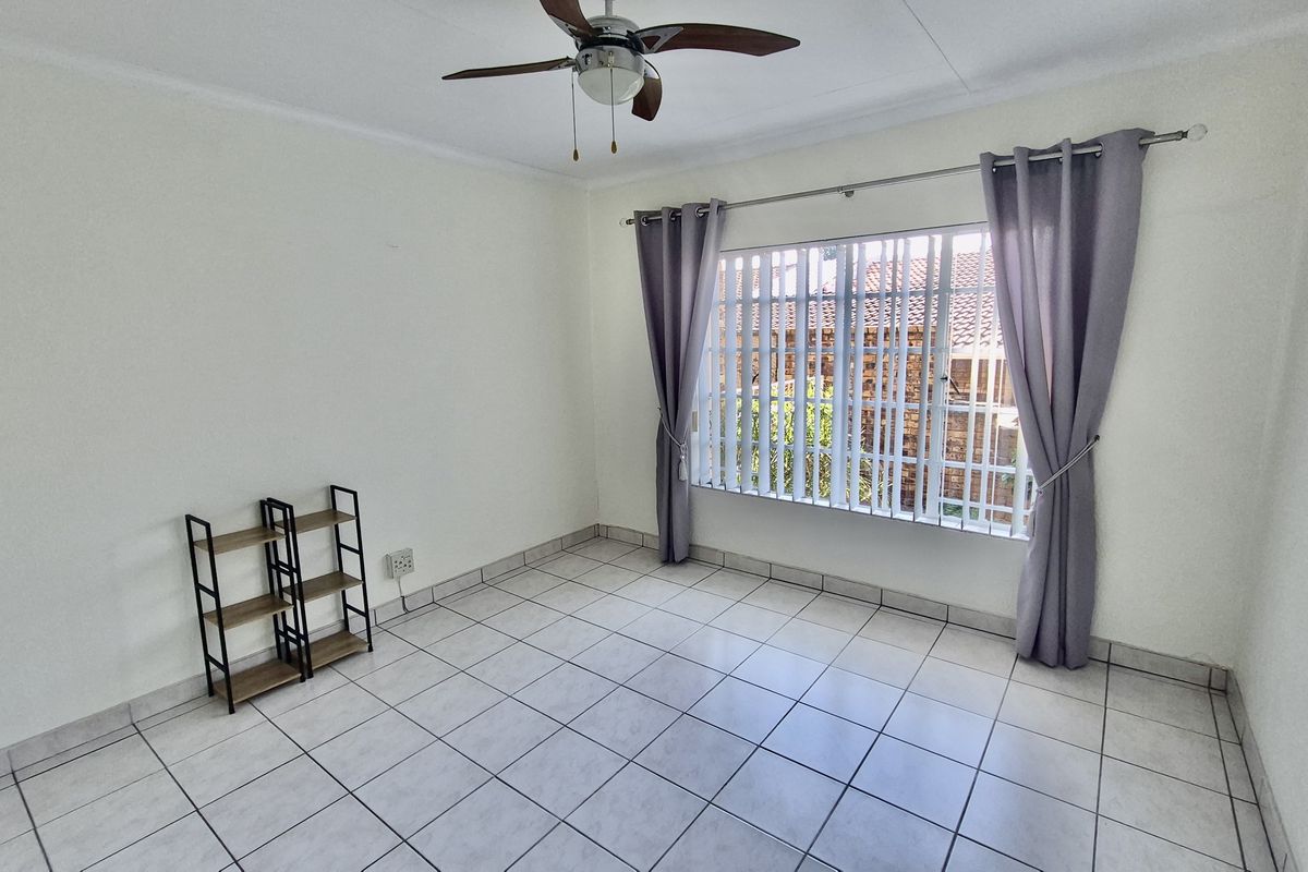 A bright, tiled bedroom featuring a ceiling fan and large window with vertical blinds, offering a clean and neutral living space.
