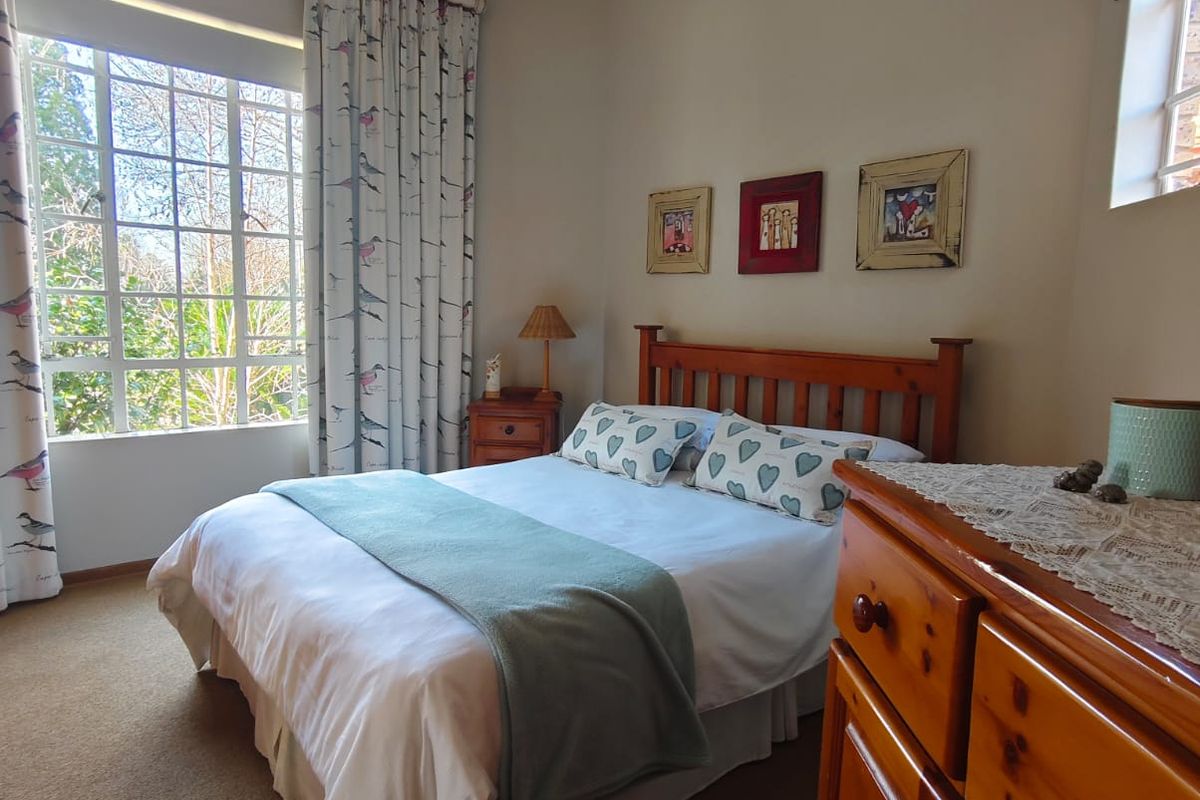 A cozy bedroom featuring large windows with natural light, neutral wall tones, and carpeted flooring.