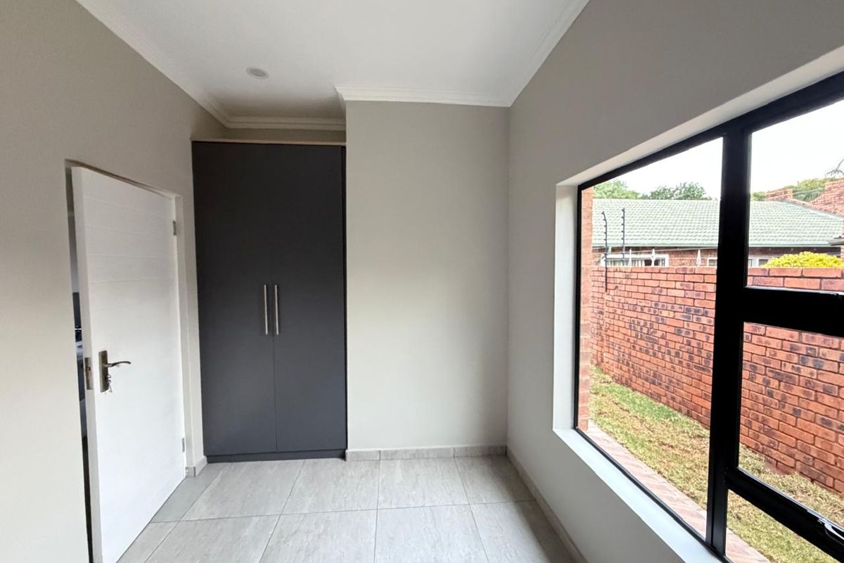 A bright, modern bedroom featuring neutral tiled flooring, a built-in dark grey wardrobe, and large windows overlooking the side garden.