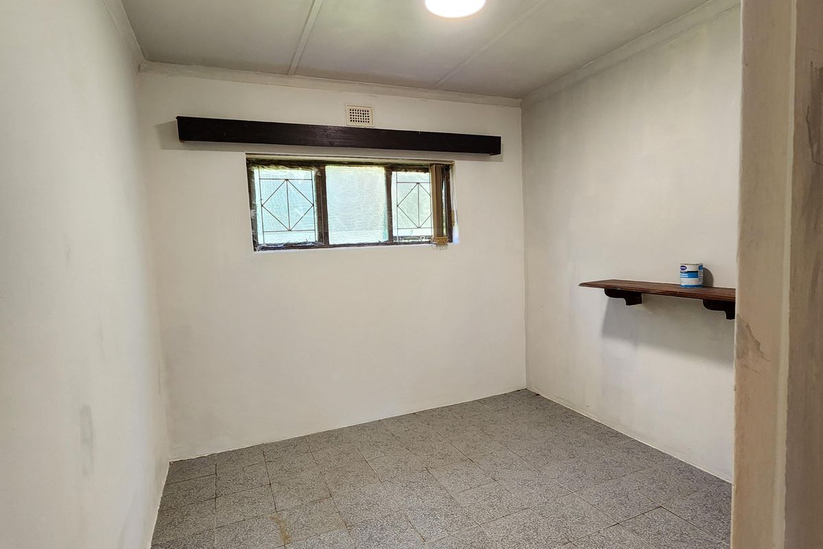 A small, empty utility room or bedroom featuring tiled flooring, a single window with security bars, and a wall-mounted shelf.