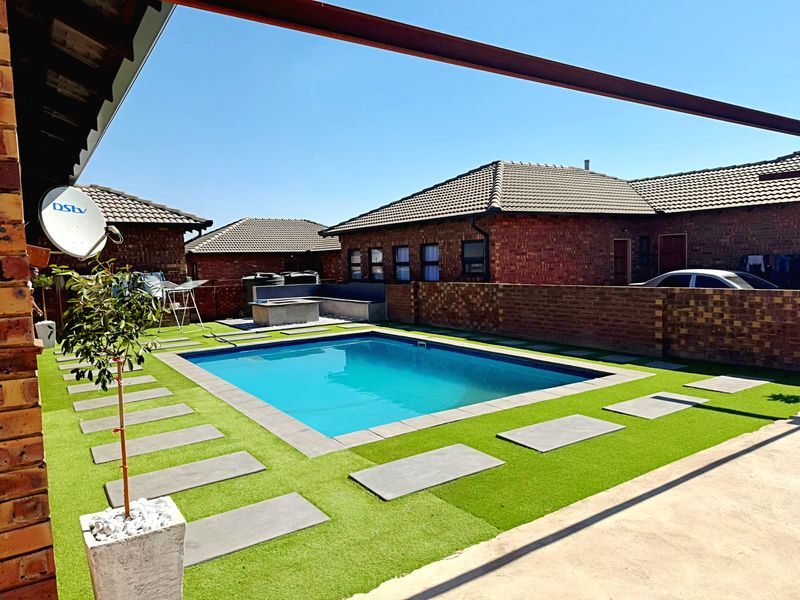 A well-maintained outdoor pool area featuring a swimming pool, artificial lawn, concrete pavers, and a brick boundary wall with built-in seating.