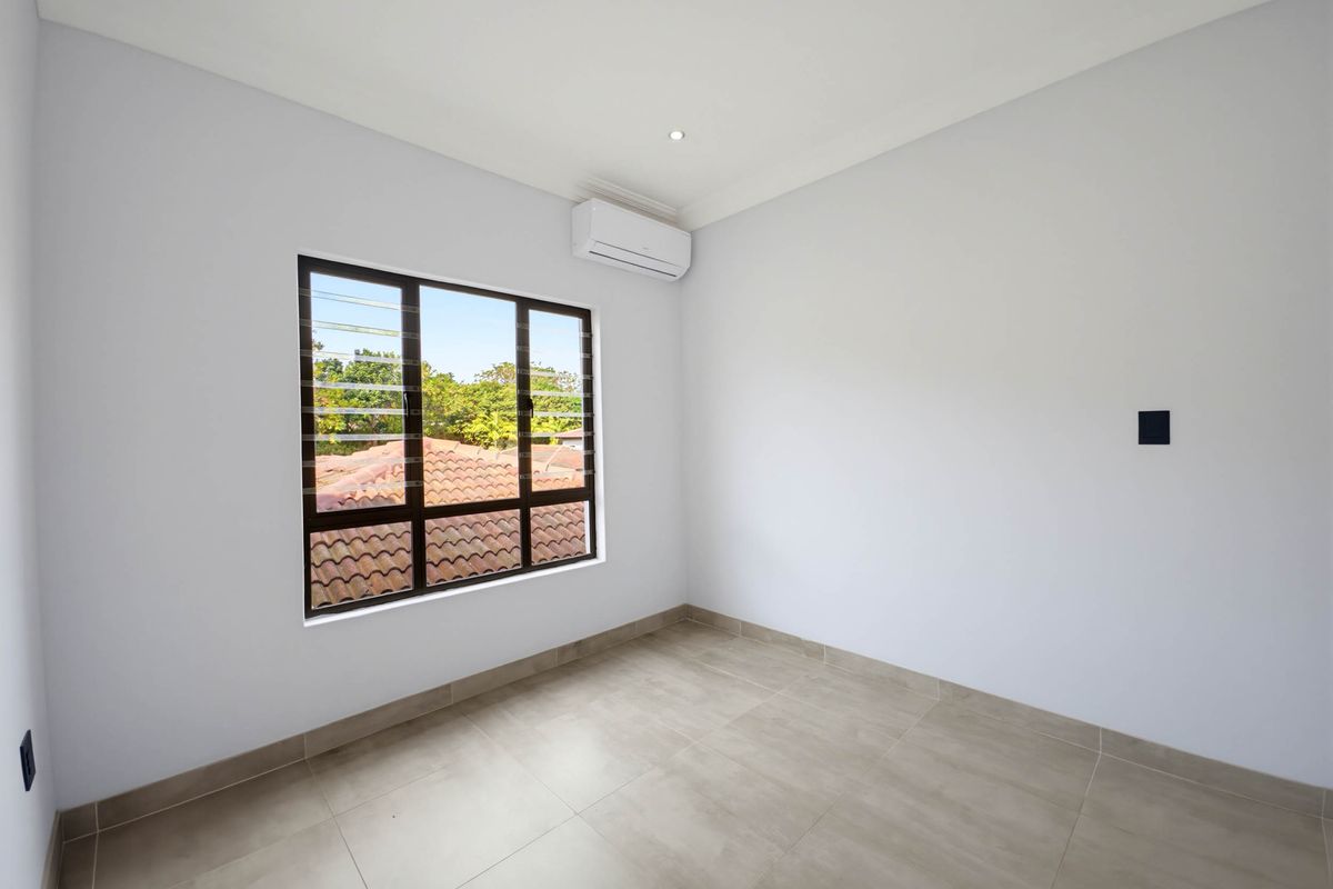 A bright, empty bedroom featuring neutral floor tiles, a wall-mounted air conditioning unit, and a window with a view of the surrounding neighborhood.