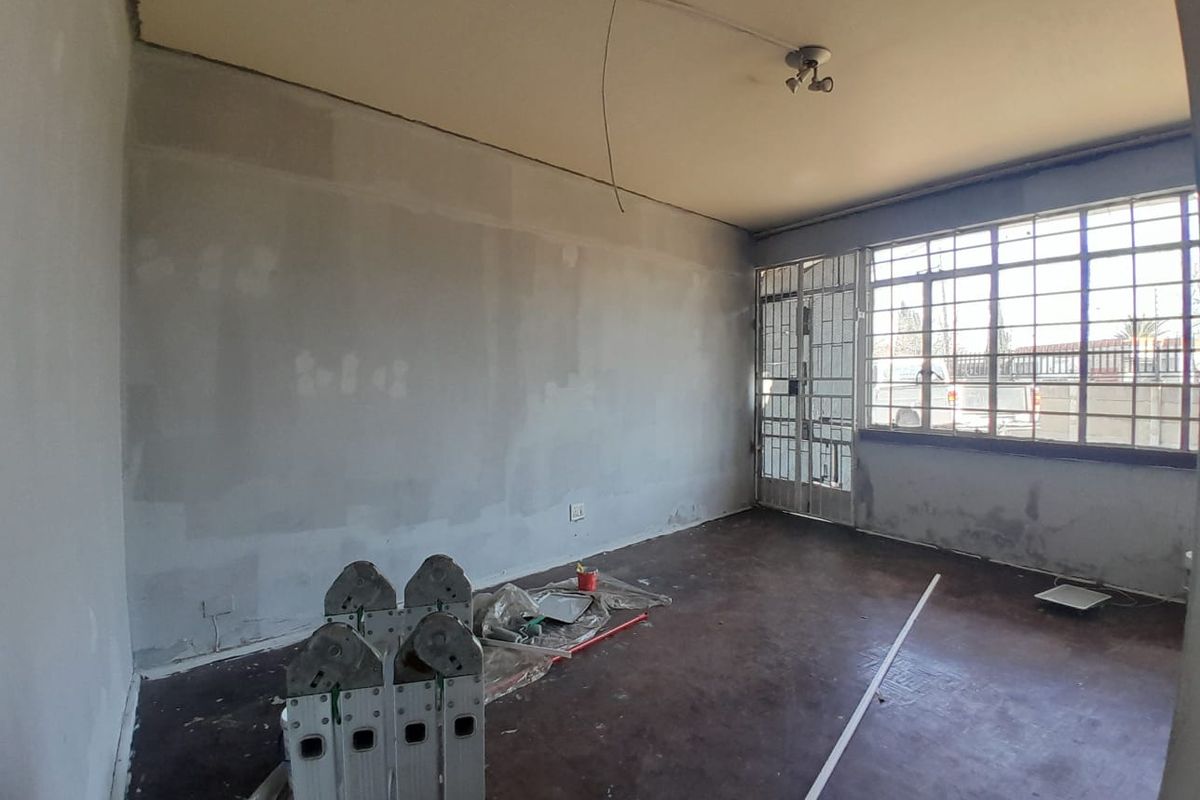 An interior view of a room currently undergoing renovation, featuring unfinished walls, exposed ceiling wiring, and a security gate at the window.