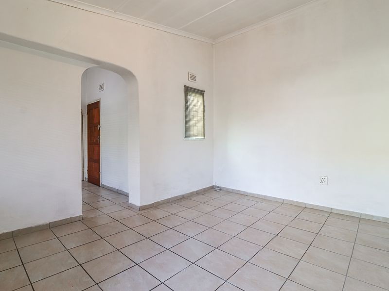 An empty, neutrally painted living area featuring tiled flooring, an arched entryway, and a small window with a fitted blind.